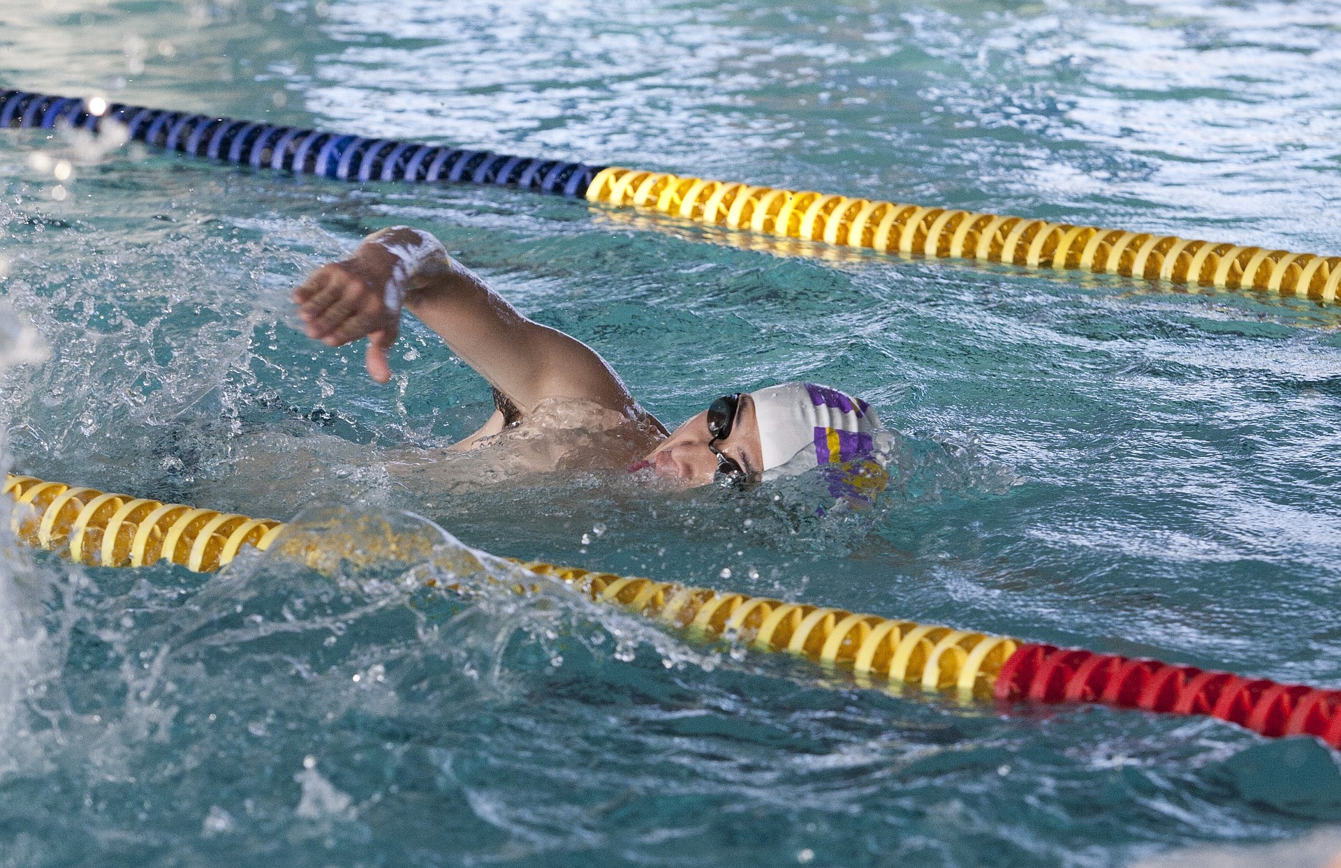 Hombre nadando en piscina olímpica. La persona tiene lentes para nadar negros y un gorro para piscina blanco con líneas moradas y amarillas a los lados.
